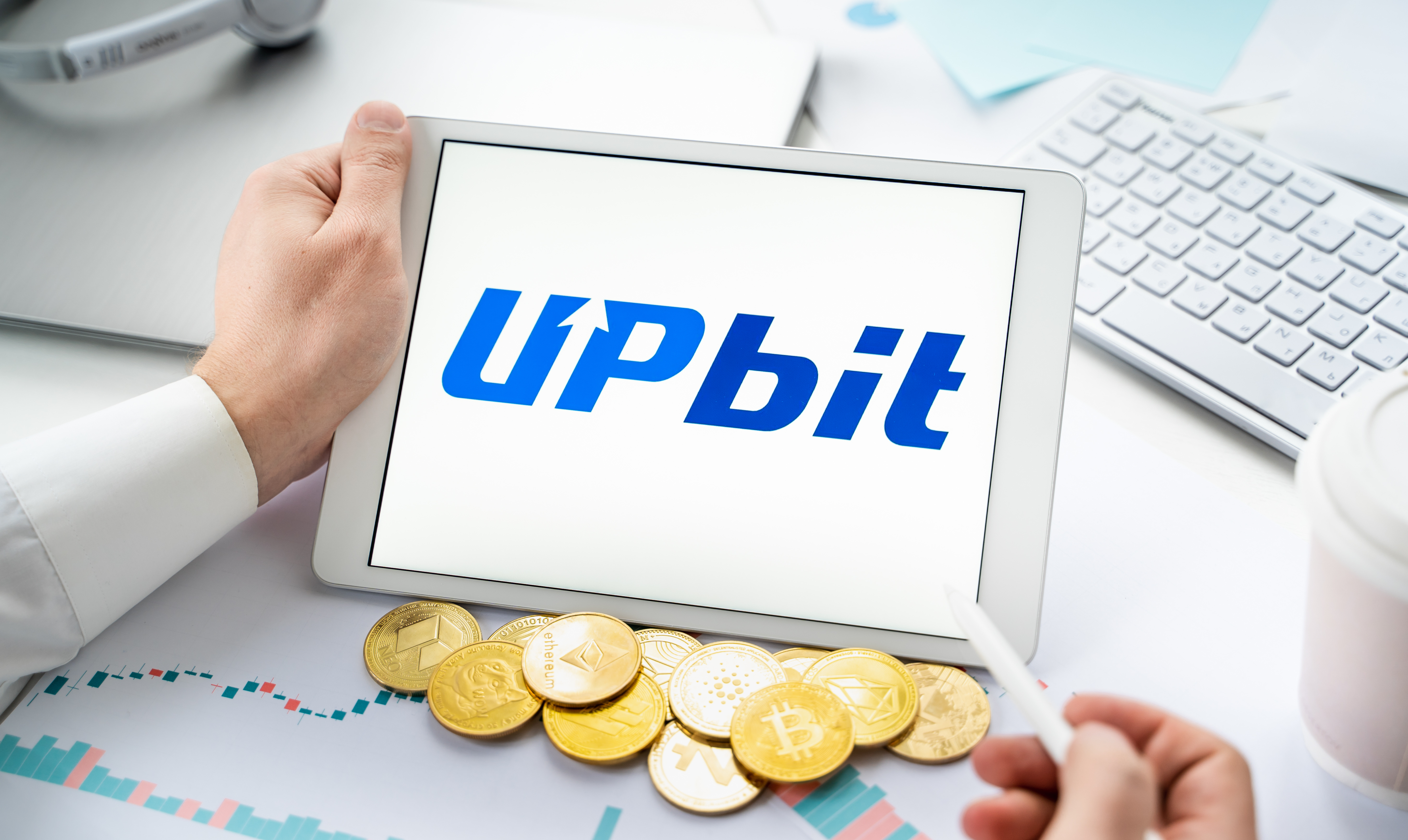 A tablet PC user looks holds a device with the Upbit logo on it. In front of the device is a pile of tokens symbolizing cryptoassets.