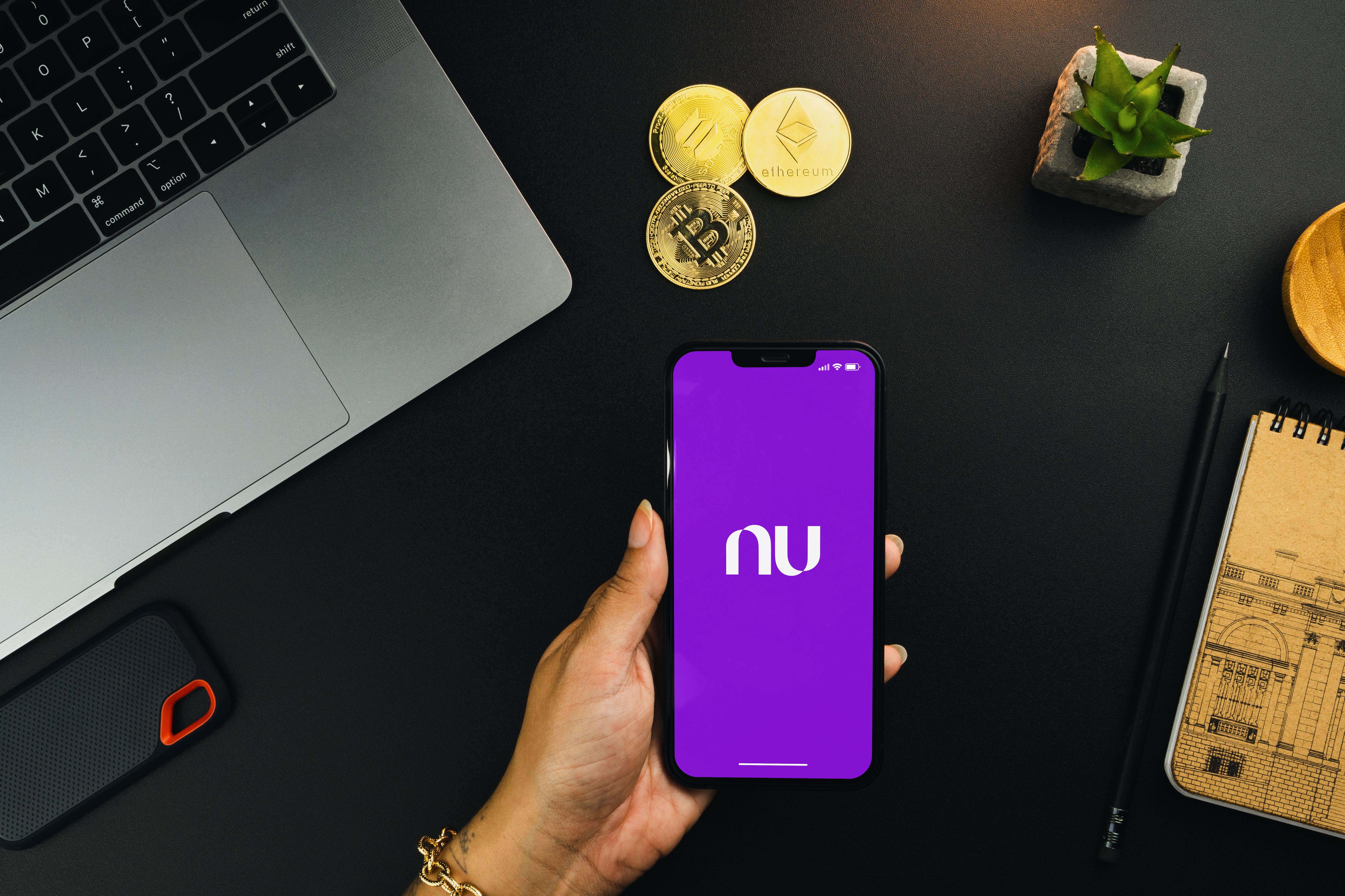 A person is holding a smartphone with the Nubank Bank app running on the screen.  On a table behind the phone, artistic representations of crypto-assets are visible, along with a laptop.