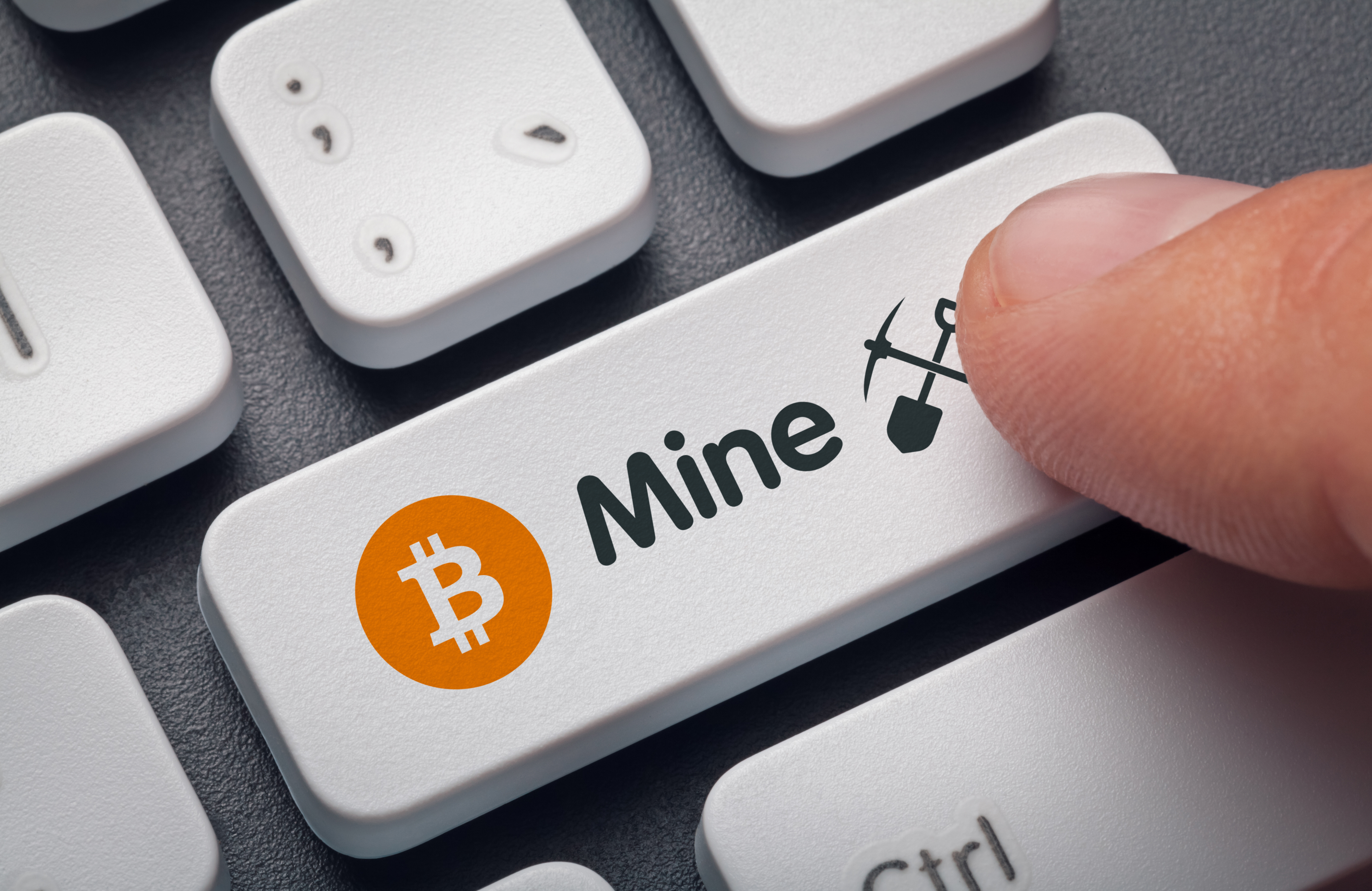 A person's finger presses a computer keyboard key.  The key is decorated with a symbol representing Bitcoin, as well as the word “mine”  and drawings of hoe and spade.