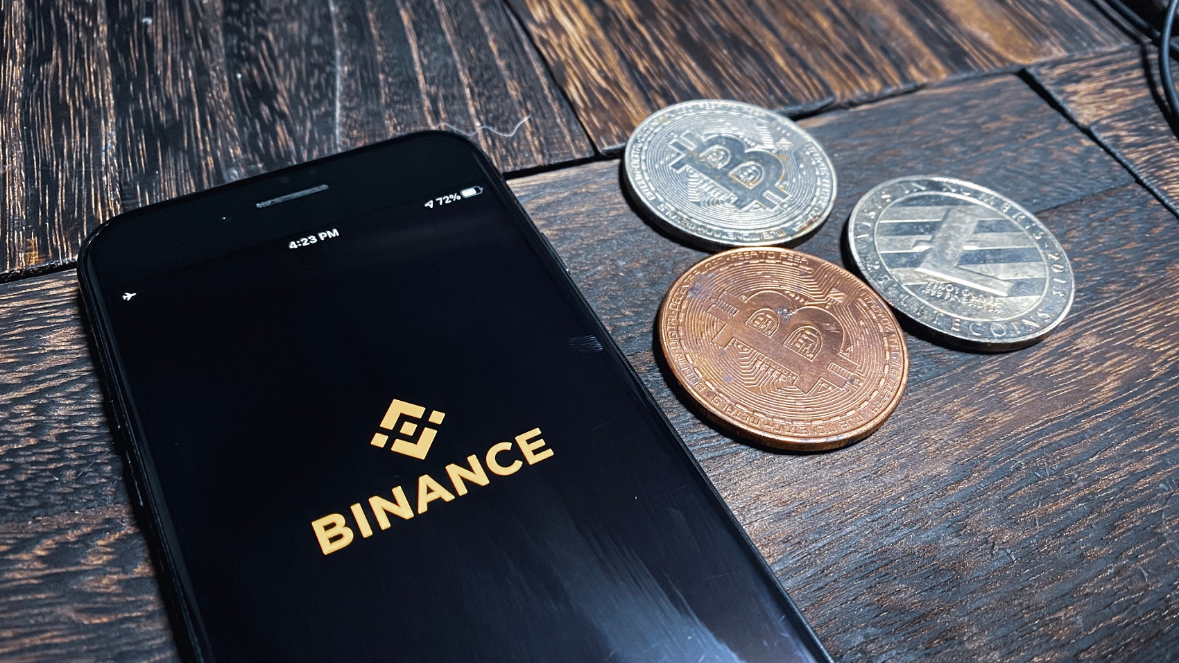 The Binance logo on the screen of a smartphone with tokens symbolizing popular cryptoassets beside it on a table.