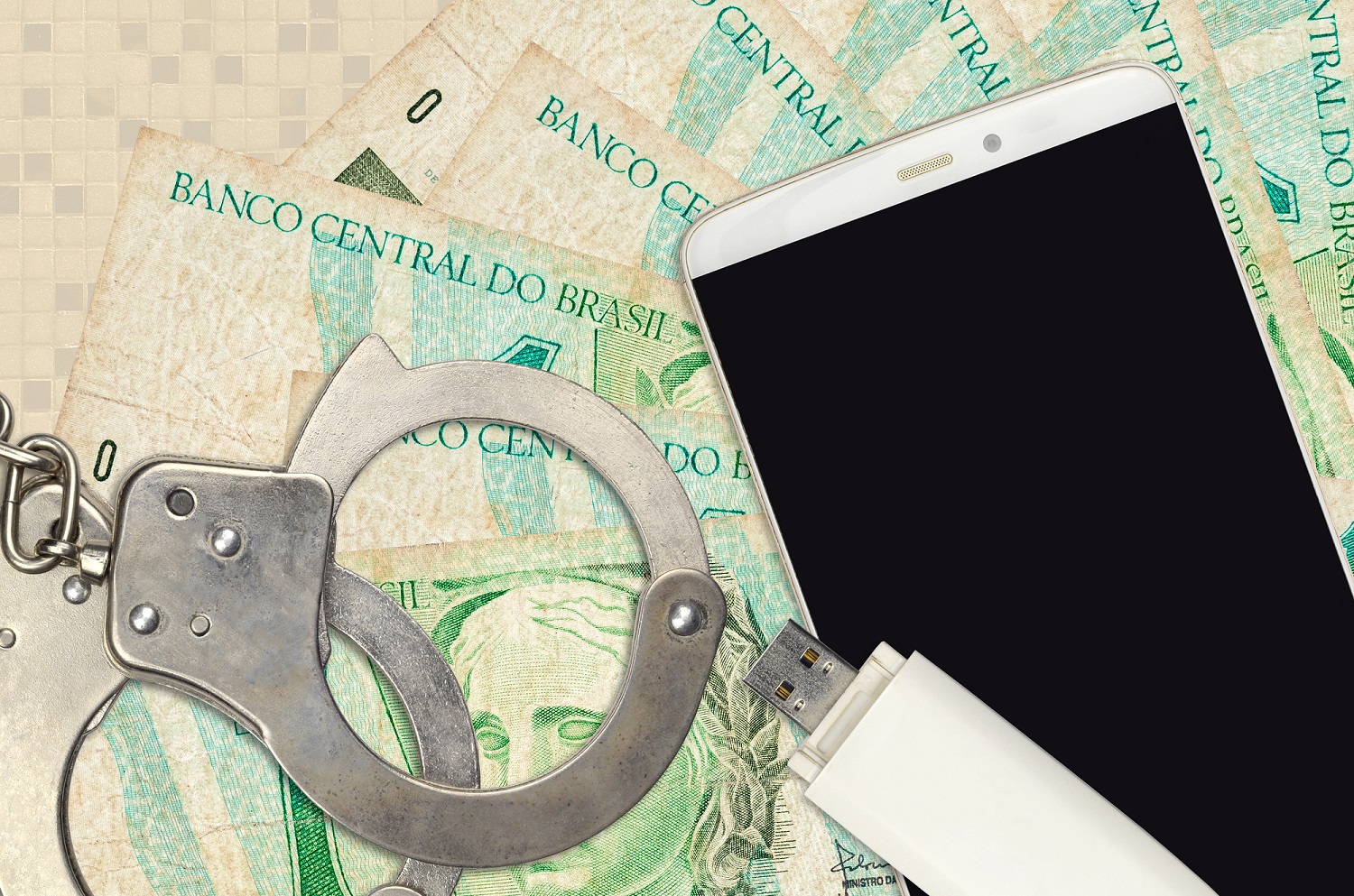 A USB storage device and a smartphone, along with a pair of police handcuffs, rest on a pile of Brazilian banknotes.