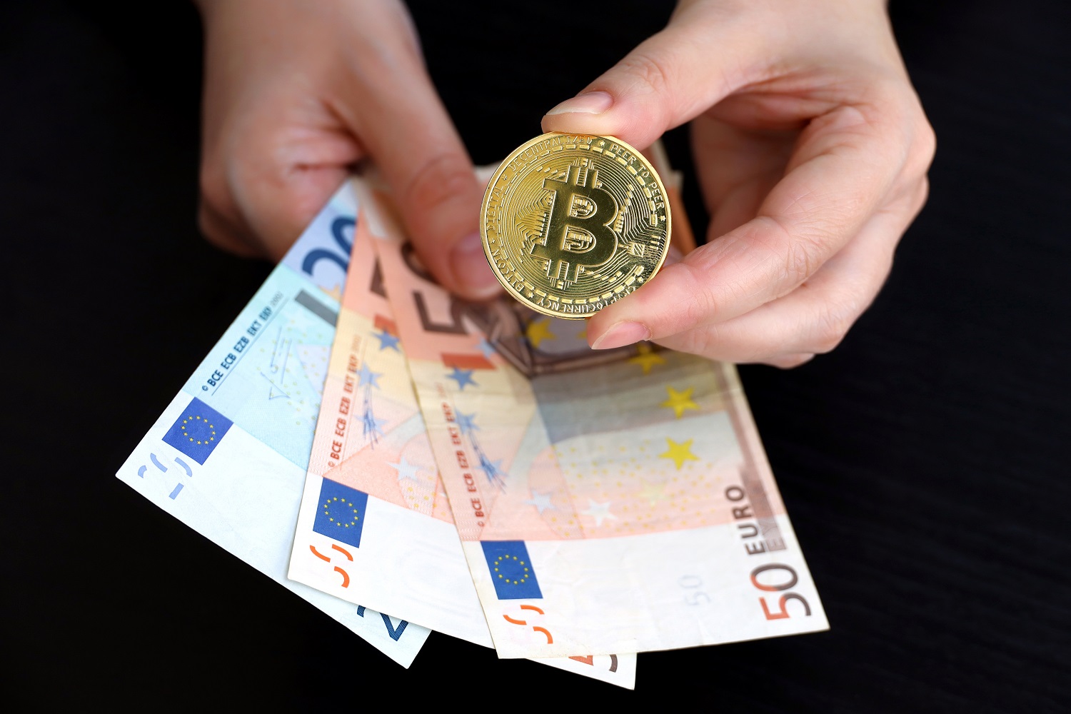 A person’s hands holding a token intended to represent Bitcoin in one hand and euro banknotes in the other.
