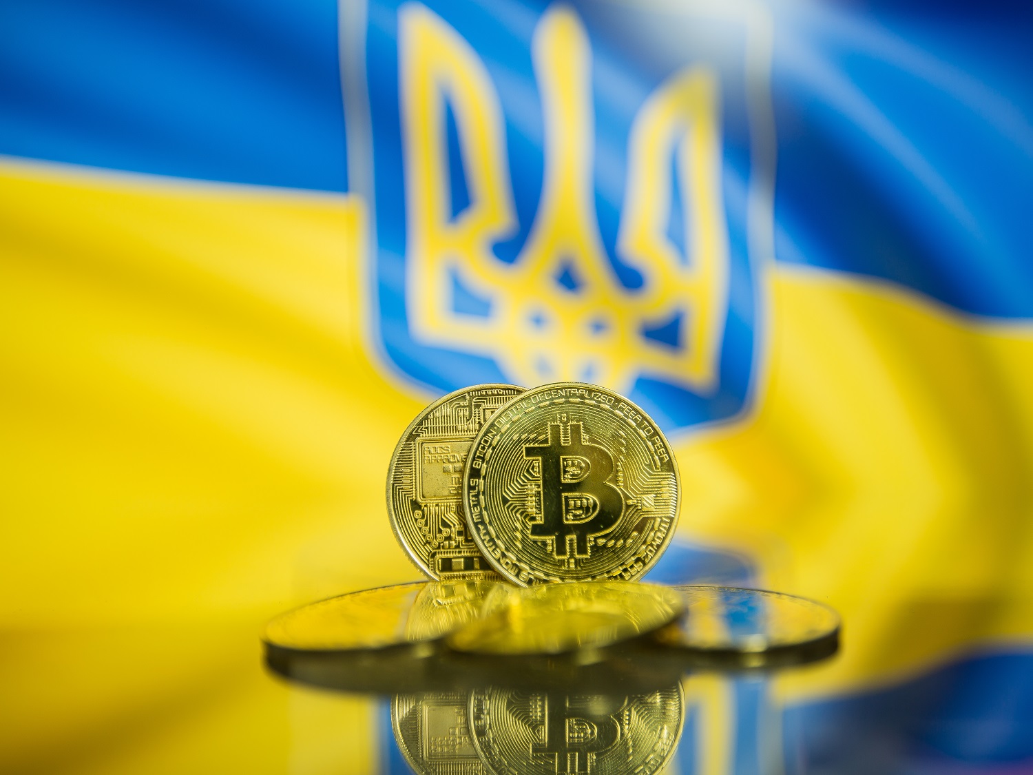Five metal coins decorated with the Bitcoin logo against the backdrop of a Ukrainian flag, featuring the trident-style national symbol.