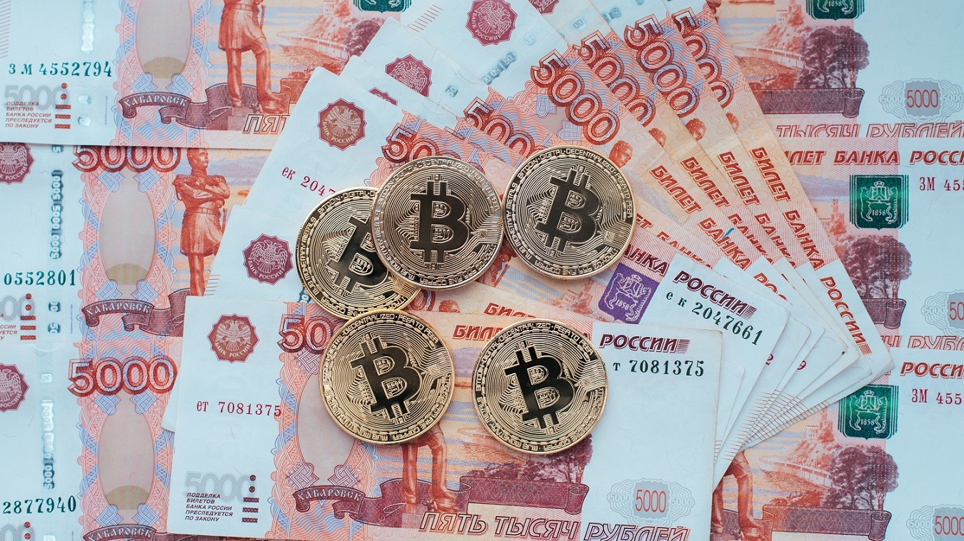 Several metal tokens intended to represent Bitcoin, rest on a pile of Russian 5000 thousand ruble banknotes on a table.