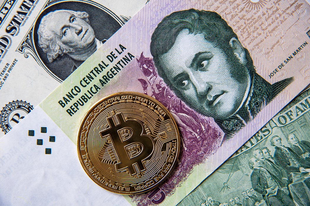 A metal token intended to represent Bitcoin on top of Argentinian peso and American dollar banknotes.