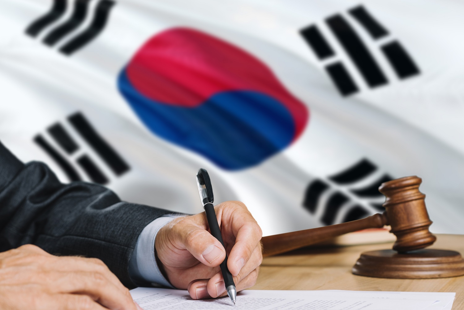 A man in a suit writes a document with a South Korean flag and a wooden gavel and block in the background.