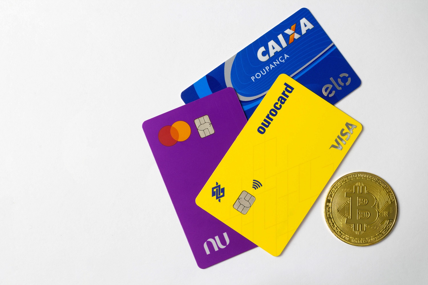 Three Brazilian bank cards next to a metal coin intended to represent Bitcoin.